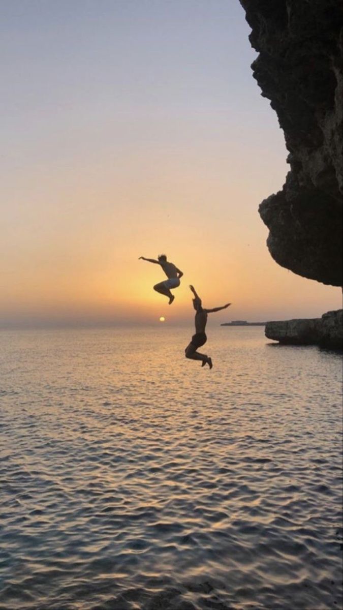 two people jumping off rocks into the ocean at sunset or dawn with one person in mid air