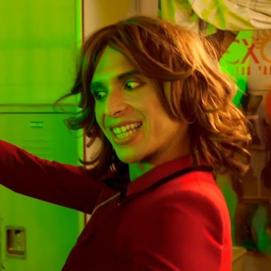 a young man with long hair and green eyes smiles while standing in front of lockers