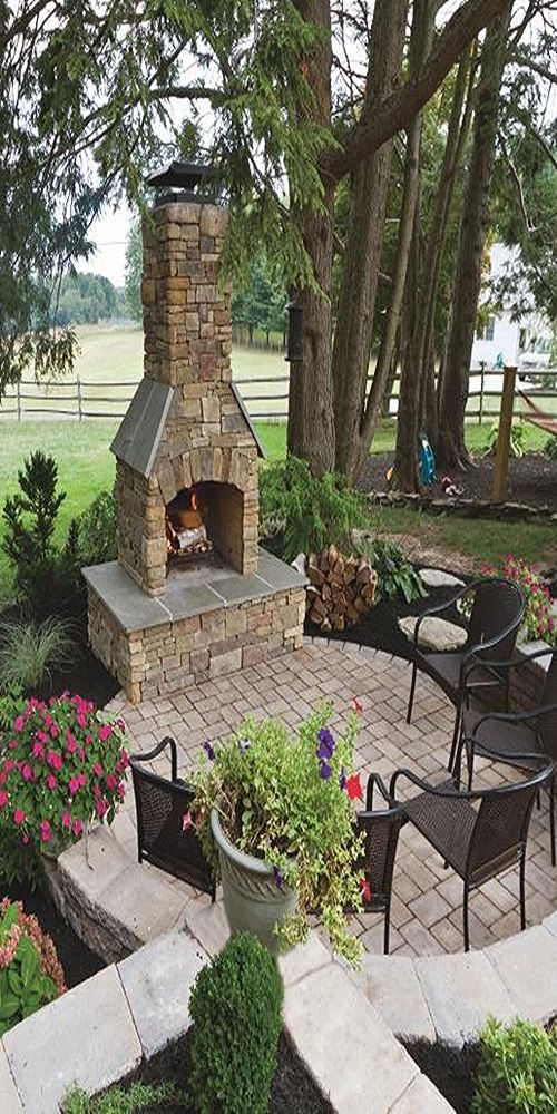 an outdoor fireplace in the middle of a patio surrounded by flowers and trees, with chairs around it