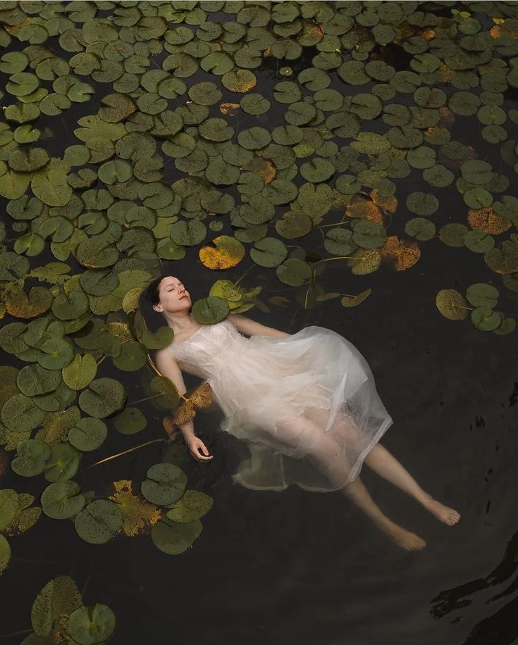 a woman floating in the water with lily pads on her back and head down, wearing a white dress