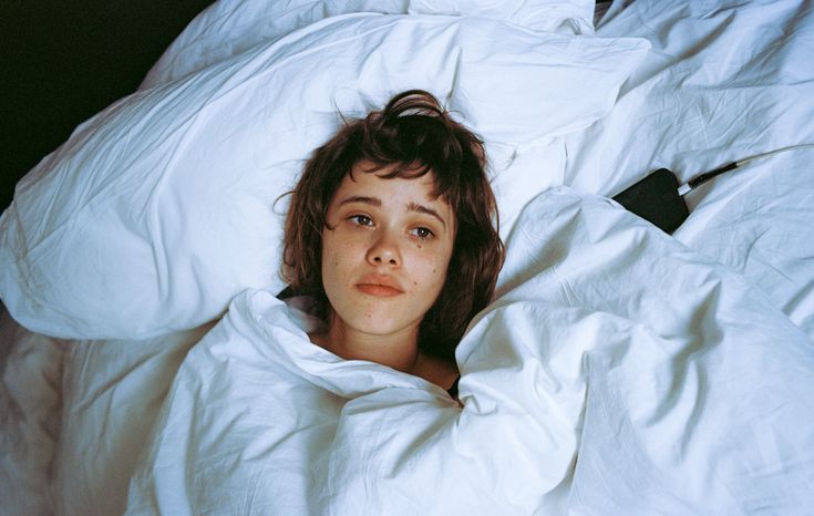 a woman laying in bed with white sheets and pillows on her head, looking up at the camera