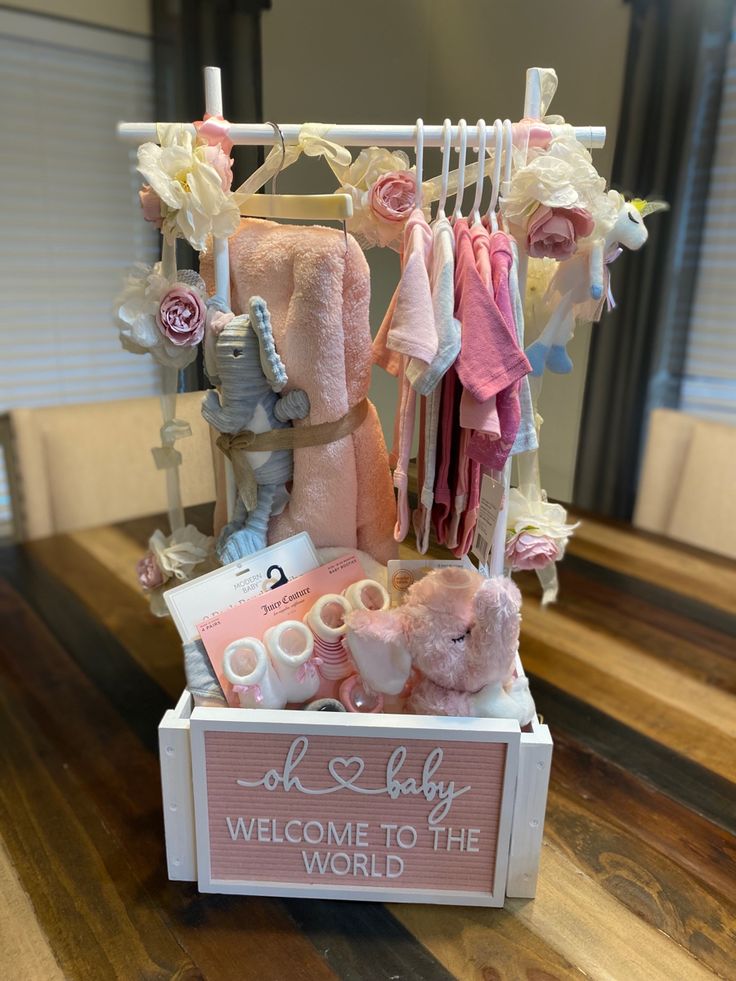 a baby welcome sign with teddy bears and clothes hanging from it's rack on a wooden table