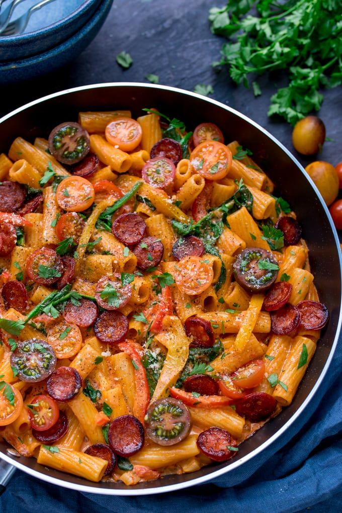 a pan filled with pasta and sausages on top of a blue cloth next to tomatoes