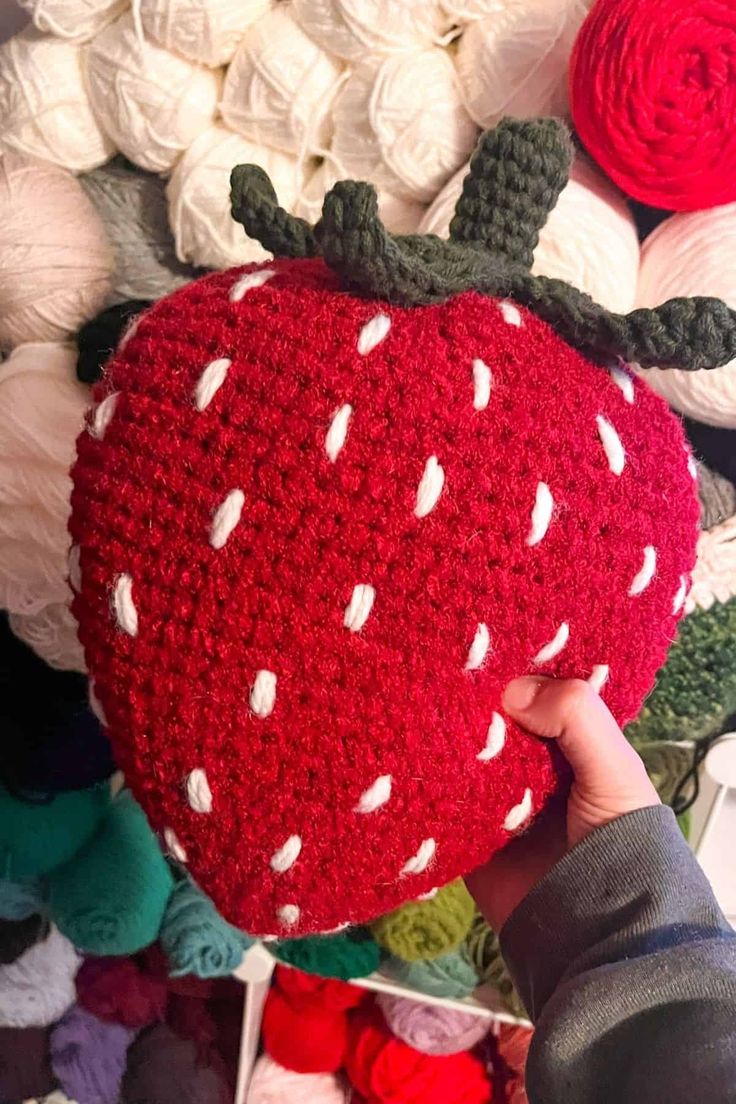 someone is holding up a crocheted strawberry cushion in front of yarn balls and needles