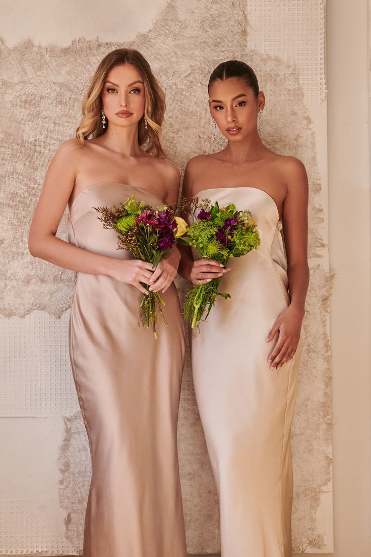 two beautiful women standing next to each other holding flowers in front of a white wall