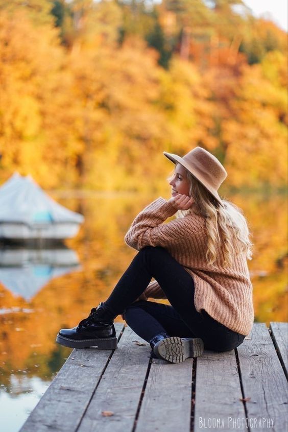 a woman sitting on a dock next to a body of water with autumn trees in the background