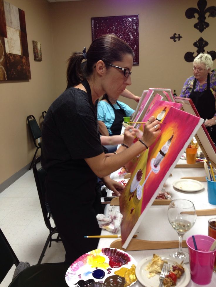 a woman is painting on an easel at a table with other people in the background