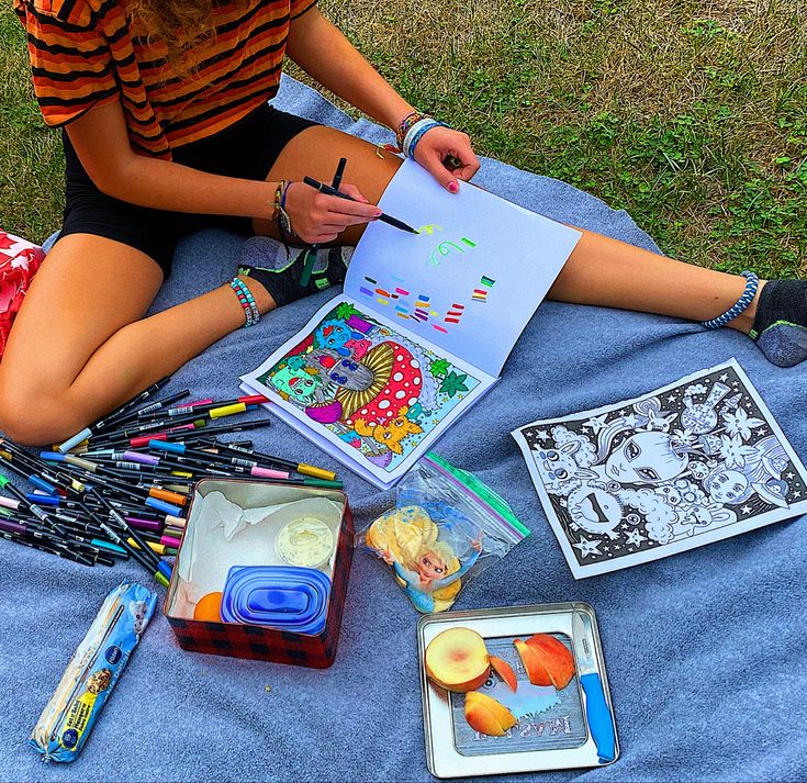 a woman is sitting on the grass with some art supplies in front of her and holding a pencil