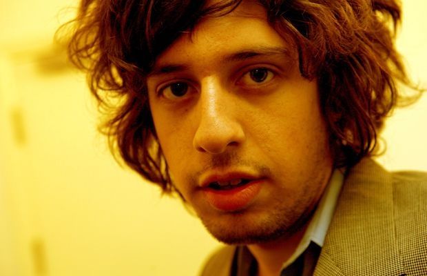 a man with long hair wearing a suit and tie looking at the camera while standing in front of a yellow wall