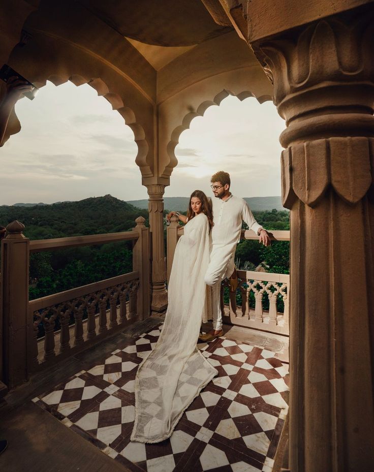 a man and woman standing on top of a balcony next to each other wearing white clothing