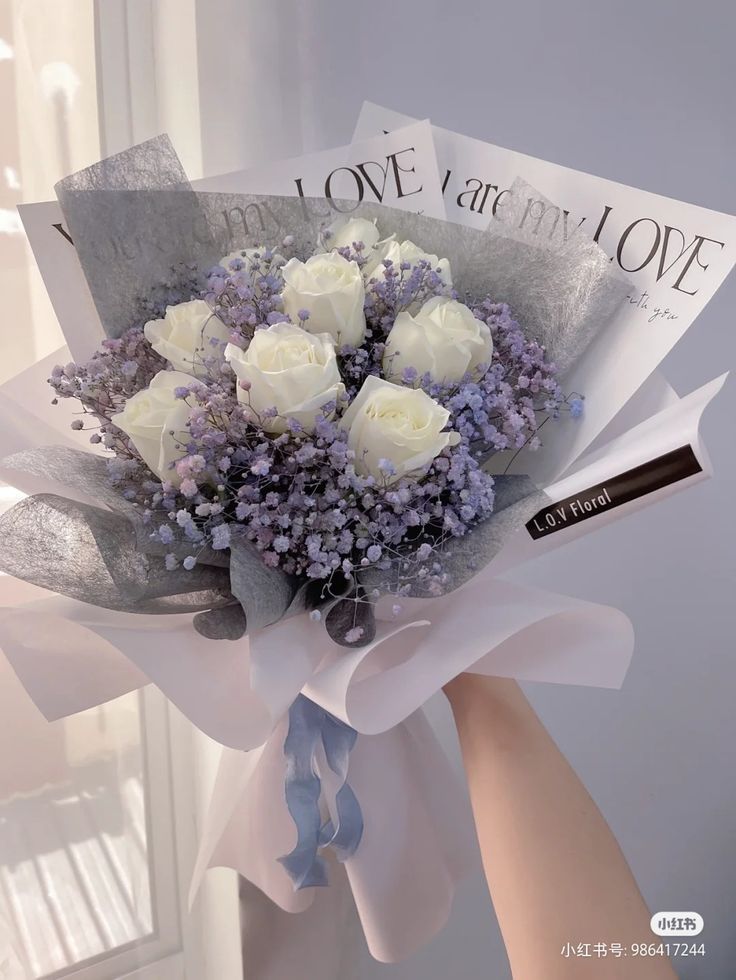 a bouquet of white roses and lavenders is held by someone's hand in front of a window