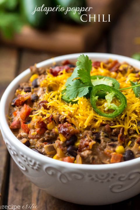 a white bowl filled with chili and cheese on top of a wooden table next to green peppers