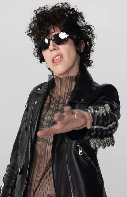 a woman in black jacket and sunglasses making a hand gesture with her right arm out