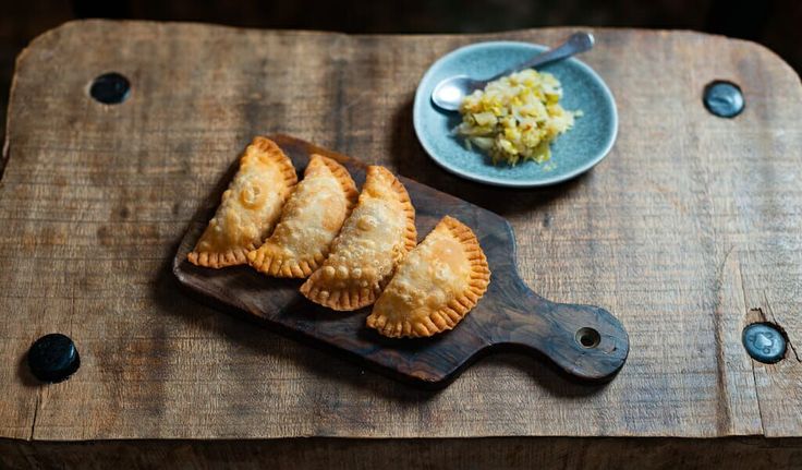 three small pastries on a wooden cutting board with a bowl of food in the background