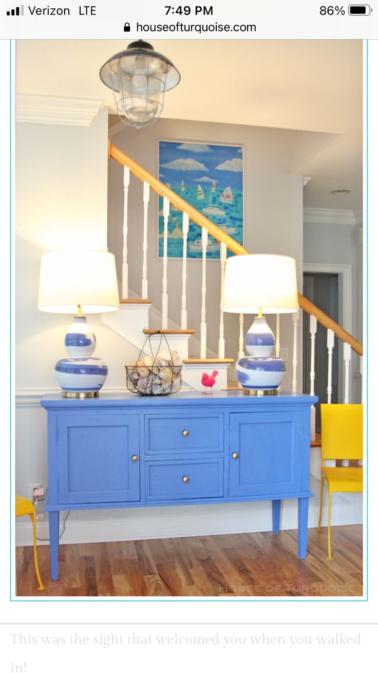 a blue dresser with two lamps on top of it next to a stair case and yellow chairs