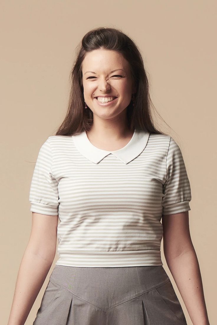 a woman is smiling and posing for the camera with her hands behind her back, in front of a beige background