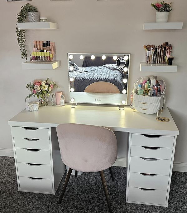 a white desk topped with a vanity mirror and lots of drawers next to a pink chair