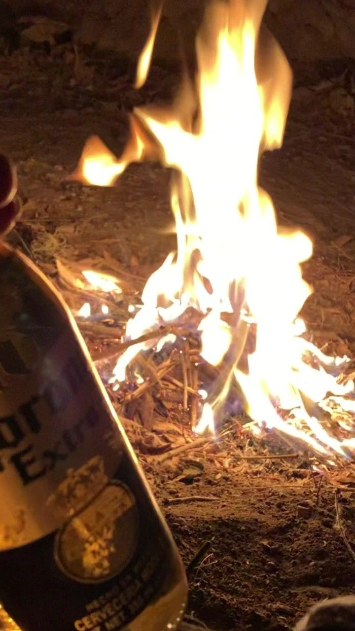 a bottle of beer sitting in front of a fire on the ground next to it