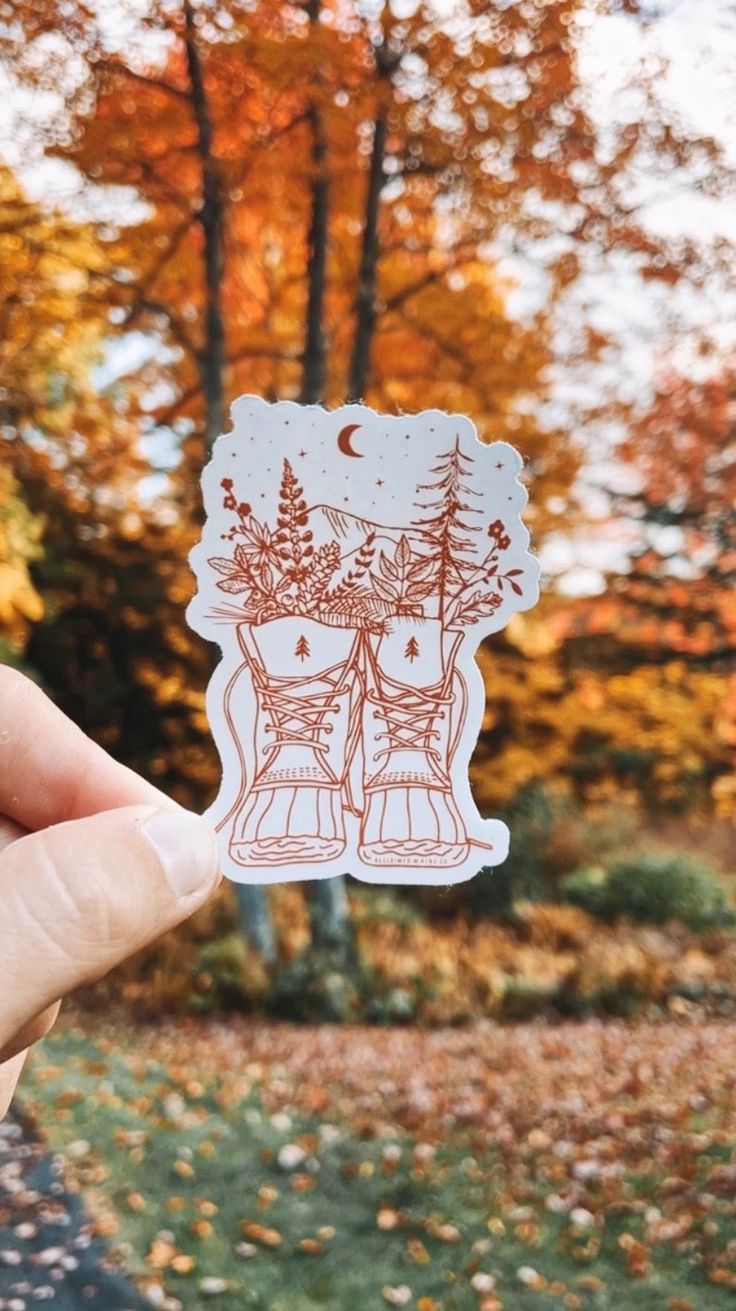 a person holding up a sticker with trees in the background and leaves on the ground