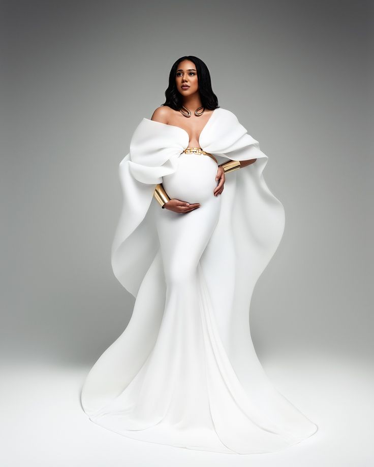 a pregnant woman wearing a white gown and gold jewelry, standing in front of a gray background