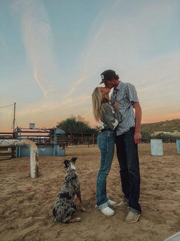 a man and woman kissing while standing next to a dog on a dirt field with an animal pen in the background