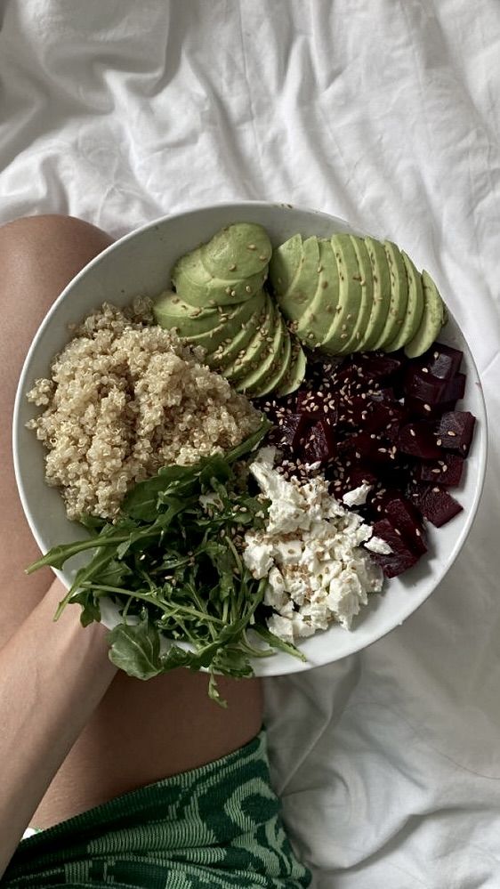 a person laying on a bed holding a white bowl filled with vegetables and avocado