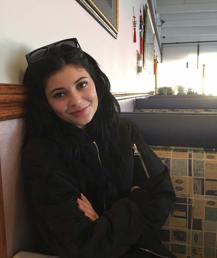 a woman with long black hair and sunglasses sitting on a bench next to a wall