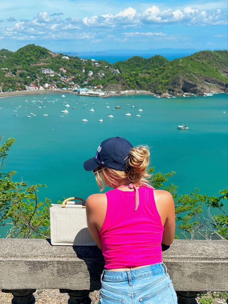 a woman sitting on a bench looking out at the ocean and boats in the water