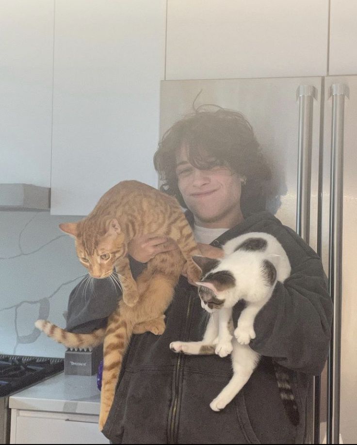 a man holding two cats in his arms while standing next to a refrigerator freezer