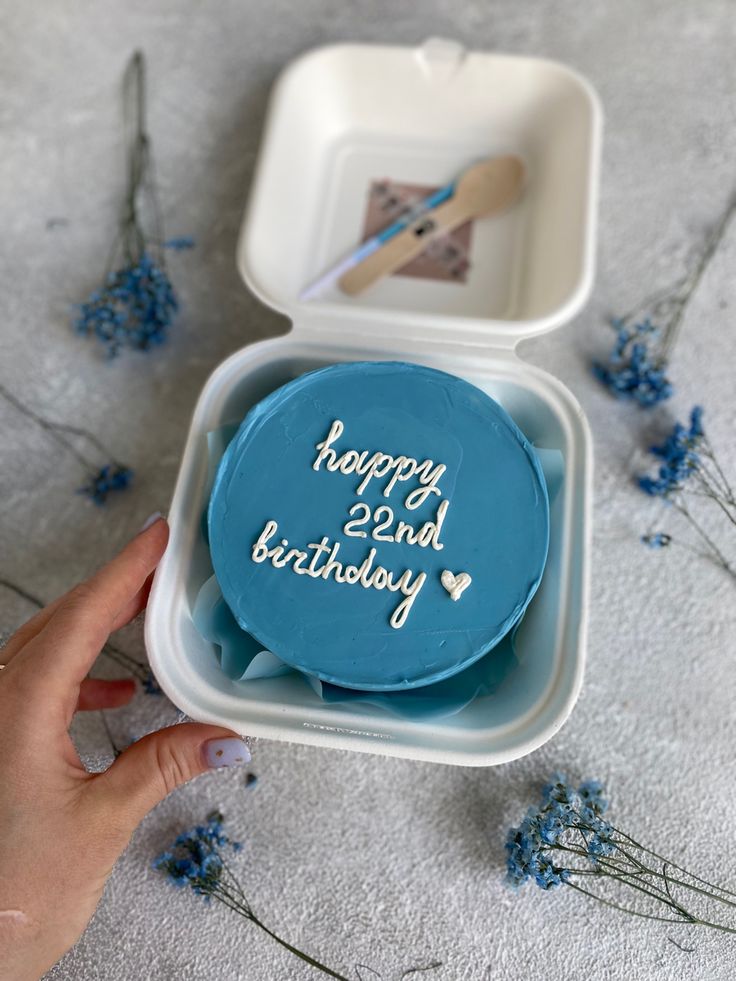 someone is holding a birthday cake in a box on the ground with blue flowers around it