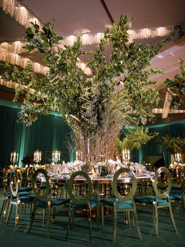 a table set up for an event with green chairs and greenery hanging from the ceiling