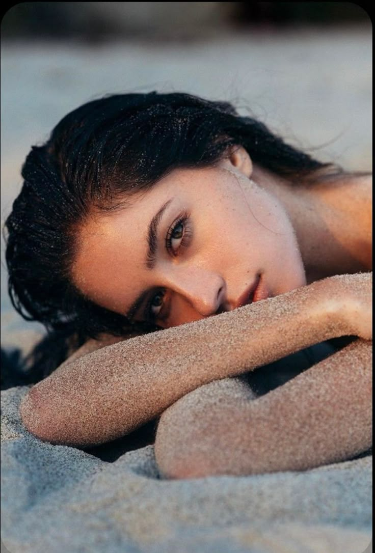 a beautiful woman laying on top of a sandy beach next to the ocean with her eyes closed