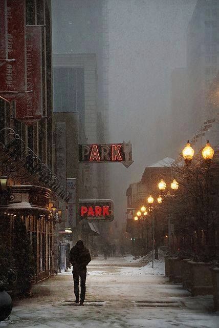 a man walking down a snow covered street in the middle of winter with lights on