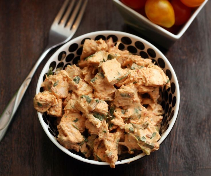 a bowl filled with chicken salad next to a plate of tomatoes and a silver fork