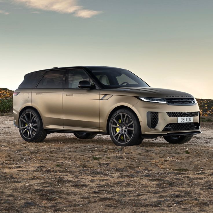 a gold range rover parked in the middle of a dirt field with grass and bushes