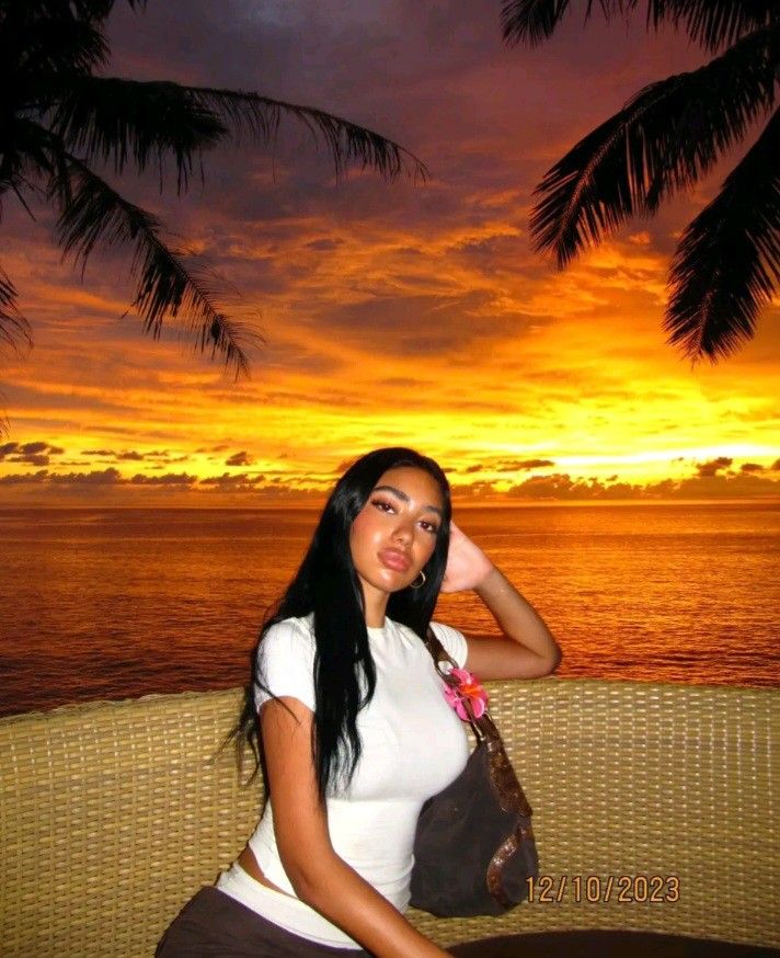 a beautiful woman sitting on top of a wooden bench next to the ocean at sunset