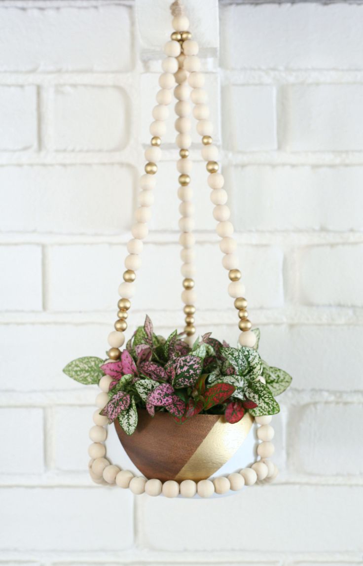 a wooden beaded hanging planter filled with flowers and greenery on a white brick wall