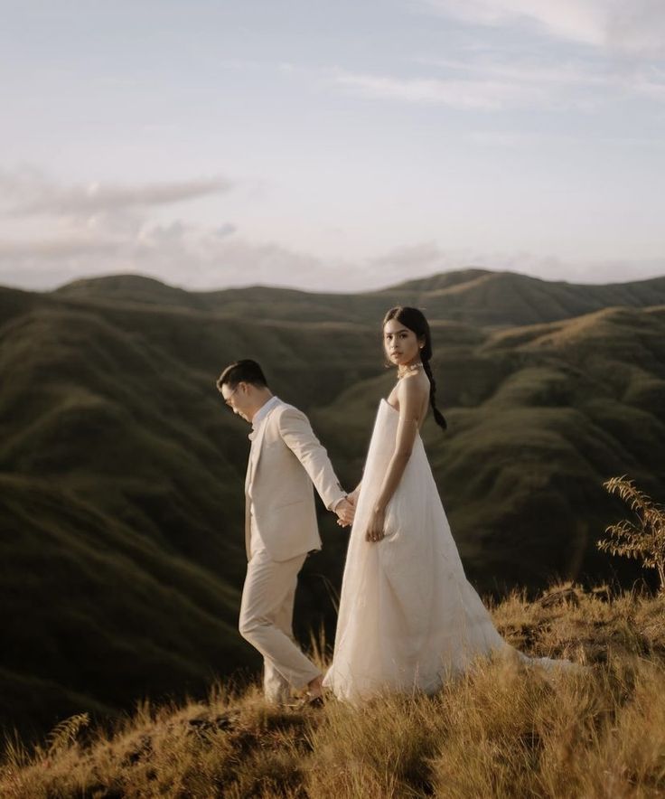 a bride and groom holding hands while standing on top of a grass covered hill with mountains in the background