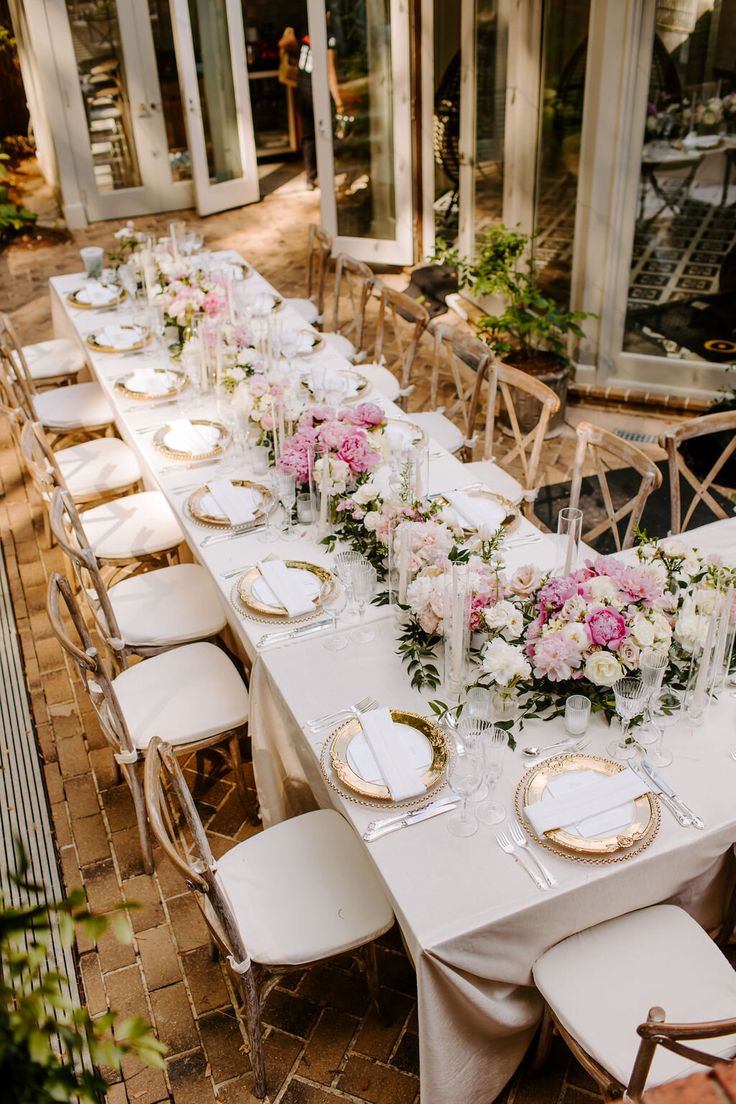 a long table is set with white and pink flowers, gold plates and silverware