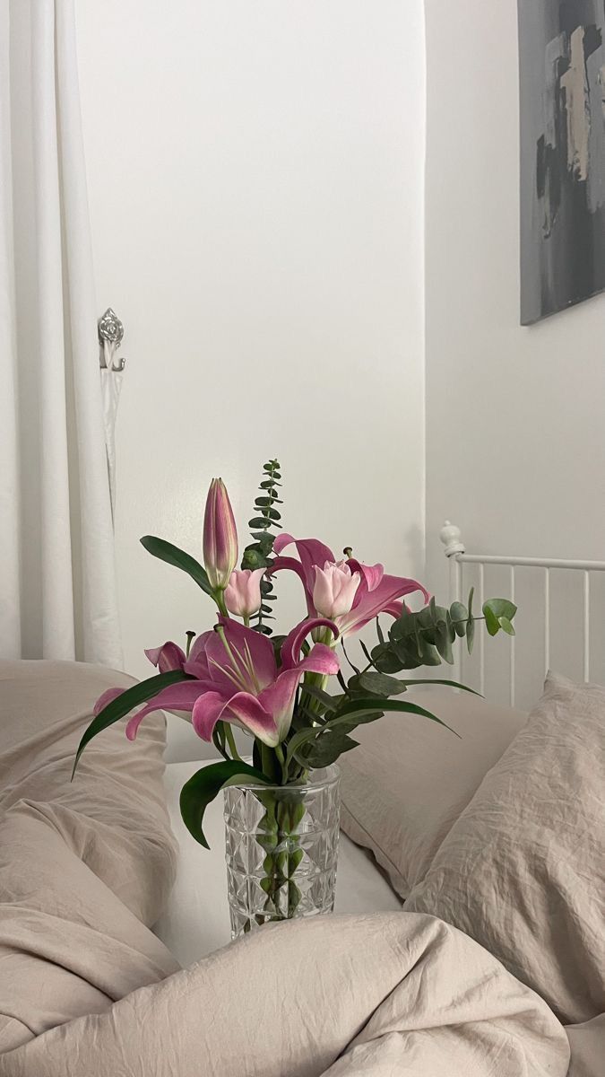 a vase filled with pink flowers sitting on top of a bed next to pillows and blankets