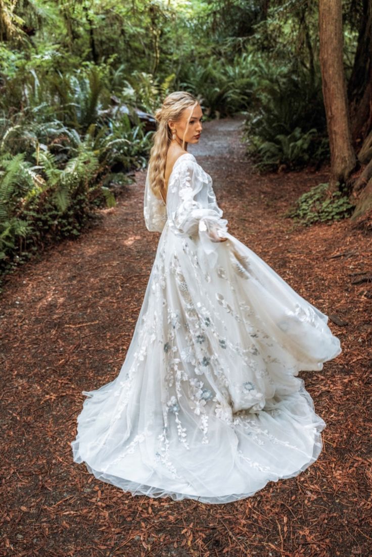 a woman in a white dress is standing on a path surrounded by trees and ferns