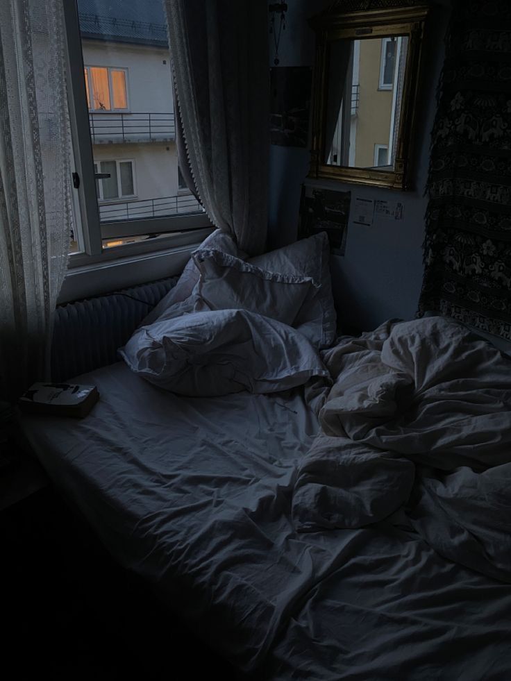 an unmade bed sitting in front of a window next to a night stand with two pillows on it