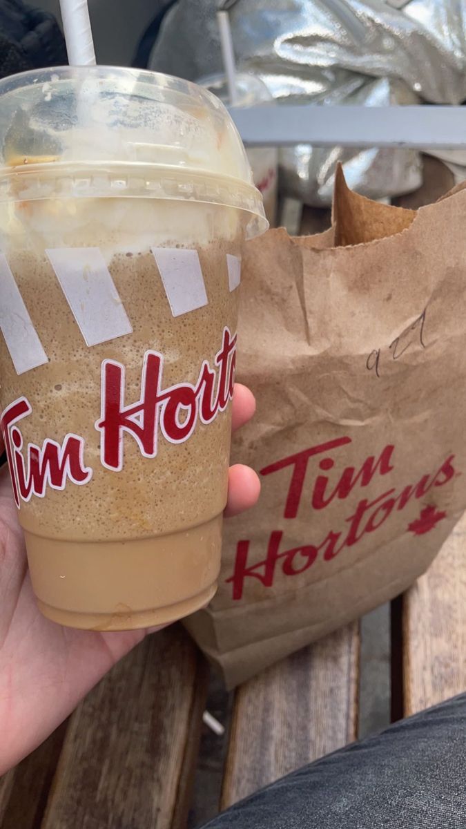 a person holding a drink in their left hand while sitting on a wooden bench next to a paper bag