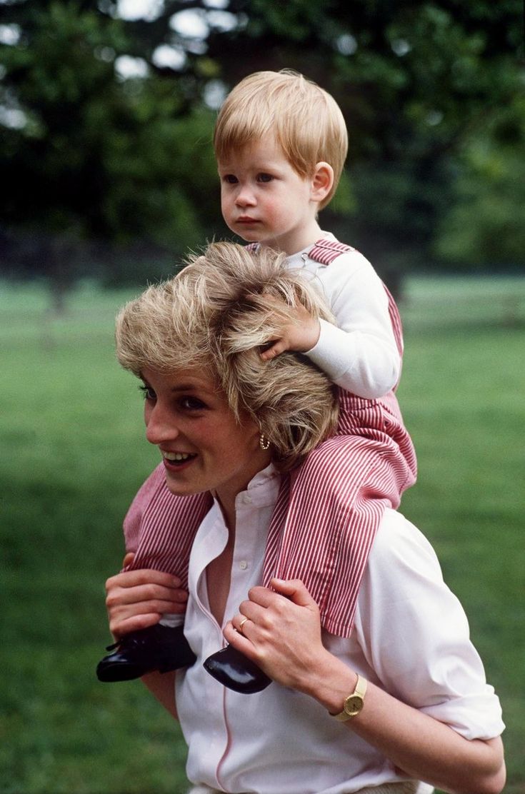 a woman carrying a child on her back in a park with trees and grass behind her