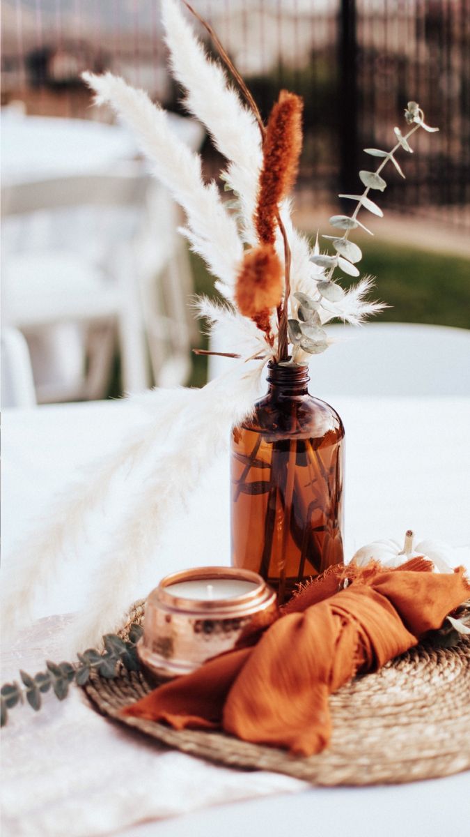 a vase with some flowers and feathers in it sitting on a table next to an orange scarf