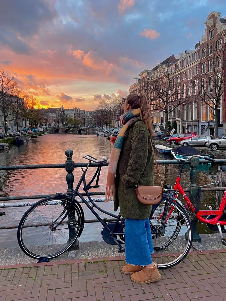 a woman standing next to a bike on the side of a river with buildings in the background