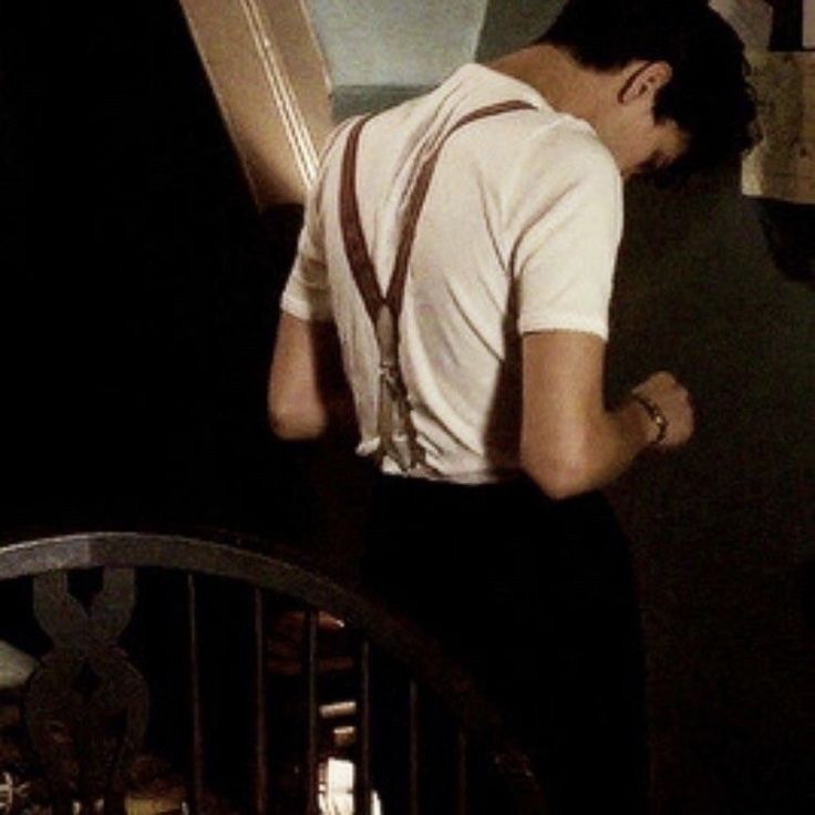 a man in white shirt and brown suspenders leaning against the wall next to stairs