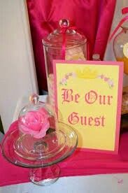 a pink table topped with a glass cake plate and a sign that says be our guest