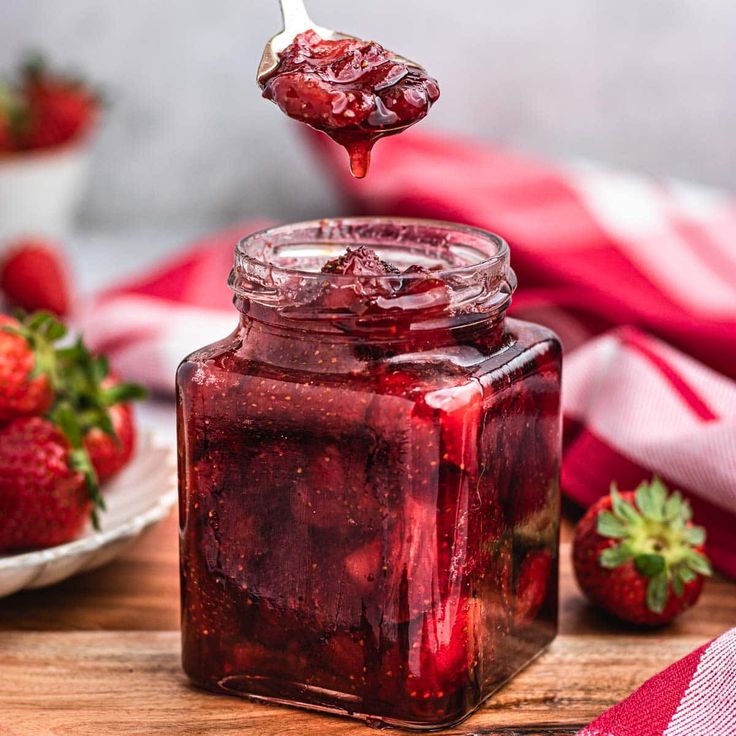 a spoon full of strawberry jam on top of a wooden table next to strawberries