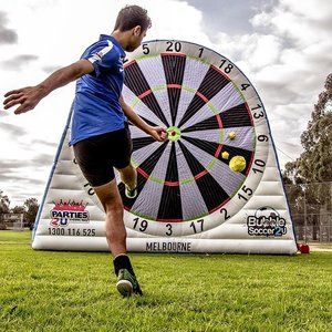 a man kicking a soccer ball in front of a giant darts board on the grass